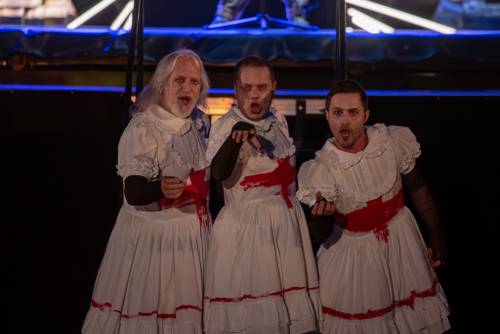 Trzech aktorów w białych sukienkach z namalowanymi czerwonymi krzyżami stoi przed sceną.