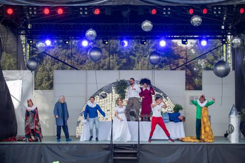 Kolorowo ubrani aktorzy stoją przed i na stołach. Tańczą.
