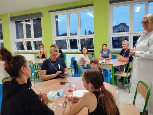 Uczestniczki podczas omówienia wprowadzenia do warsztatów siedzą przy stolikach. Prowadząca w białym fartuchu stoi.