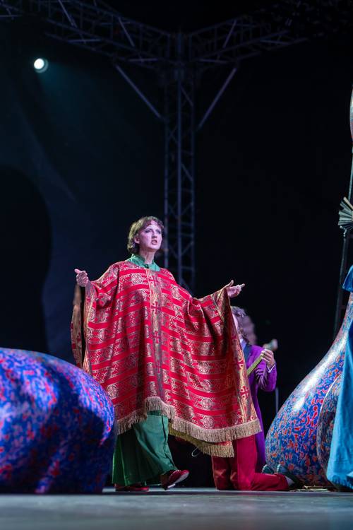 Kobieta z rozłożonymi rękami odziana w koc kroczy przez scenę.