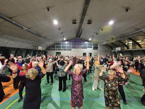 Seniorzy tańczą z uniesionymi rękami. Widać dużą grupę ludzi.