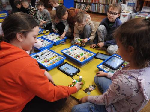 Grupa dzieci siedząca na macie z zestawami kloców i tabletami. Widok zza pleców.