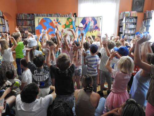 Przedstawienie w bibliotece, dzieci stoją z rękami w górze, przed nimi Smerf