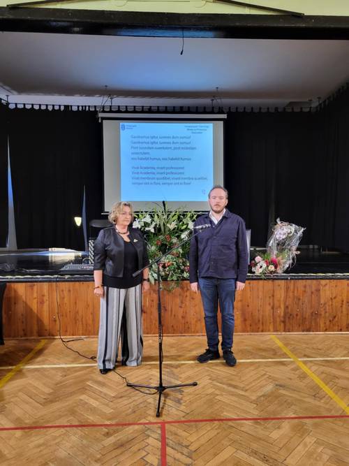 Na zdjęciu koordynatorka UTW, obok stoi mężczyzna, z tyłu na wyświetlaczu widać słowa pieśni Gaudeamus igitur