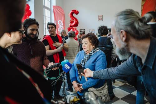 Kobieta w niebieskim swetrze i szaliku udziela wywiadu, otoczona przez kilku mężczyzn z mikrofonami.