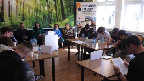 Uczniowie siedzą przy stolikach i czytają to co jest napisane na kartkach