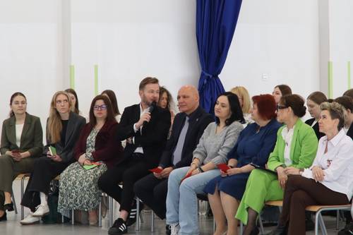 Jeden z gości siedzący wśród innych trzyma w ręku mikrofon, do którego mówi