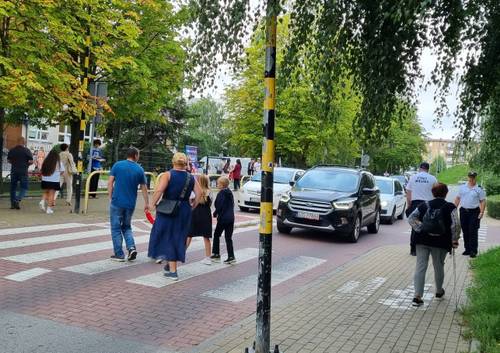 Strażnik miejski stoi na chodniku przy przejściu dla pieszych, w pobliżu którego przechodzą dzieci.