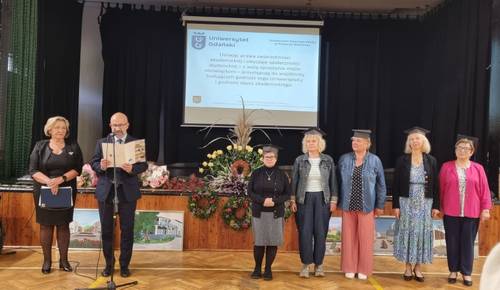 Przed sceną stoi grupa osób, nowi studenci Uniwersytetu Trzeciego Wieku i koordynatorzy
