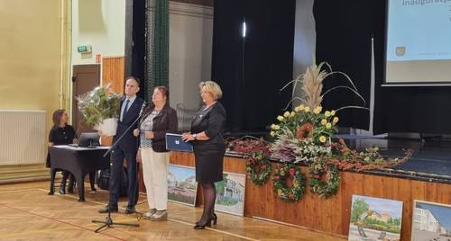 Dwie kobiety i mężczyzna stoją przed podestem przy którym stoją obrazy i kwiaty