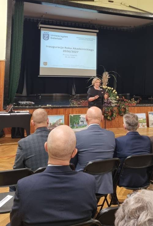 Grupa mężczyzn siedzi na krzesłach, przed nimi stoi kobieta z teczką w ręku