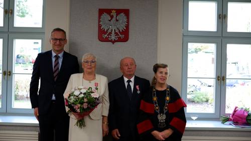 Dwóch panów i dwie panie stoją przy ścianie, na której wisi godło