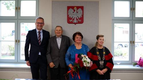 Dwóch panów i dwie panie stoją przy ścianie, na której wisi godło