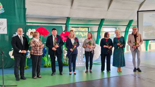 Grupa osób stoi na zielonym tle, część z nich trzyma w dłoniach kwiatka