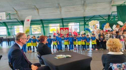 Zbliżenie na stolik przy którym siedzą dwie pani i jeden pan, w tle grupa dzieci w żółto-niebieskich strojach