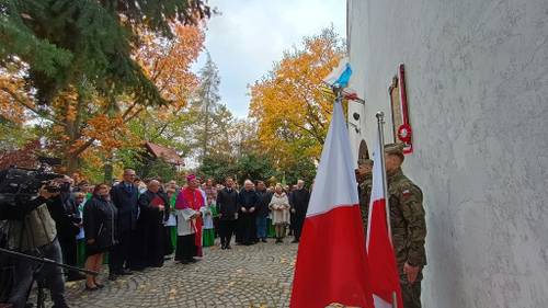 Widok na żołnierzy stojących za flagami i grupę ludzi stojących w półokręgu przed nimi