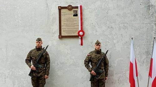 Dwóch żołnierzy stoi na białej ścianie, na której wisi tablica z biało-czerwoną wstążką