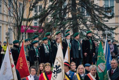Uczestnicy ze sztandarami, w tle harcerze z zielonymi nakryciami głowy.